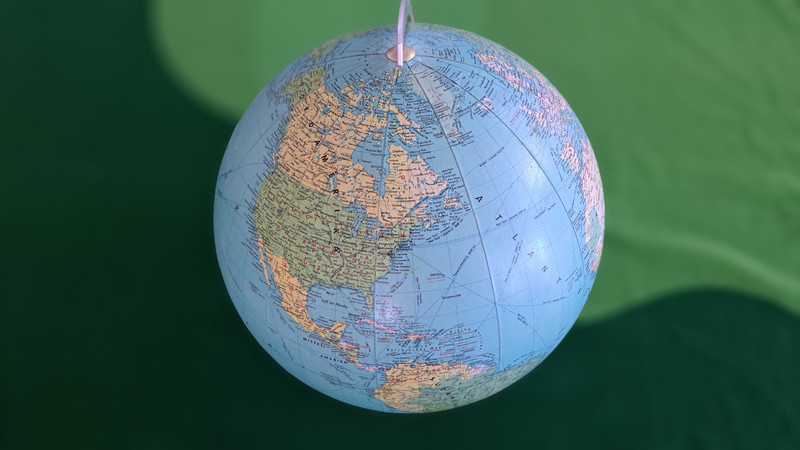 Auf dem Bild ist ein Globus zu sehen, der die Erde darstellt. Der Globus hängt an einer Schnur und zeigt vor allem den amerikanischen Kontinent, einschließlich Nord- und Südamerika. Der Hintergrund ist grün, vermutlich eine Wand oder ein Vorhang. Der Globus selbst ist detailliert mit geografischen Merkmalen und Ländergrenzen versehen.