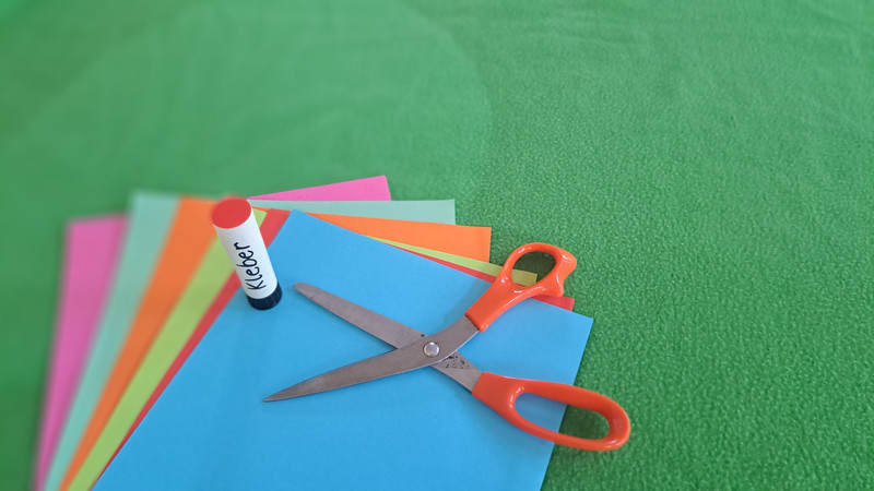 Auf dem Bild sind verschiedene Bastelmaterialien zu sehen. Es gibt mehrere bunte Papierbögen in den Farben Rosa, Rot, Gelb, Grün und Blau. Auf diesen Papierbögen liegen eine Schere mit orangefarbenen Griffen und ein Klebestift.