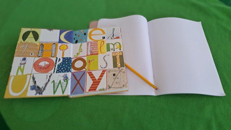 Auf dem Bild ist ein aufgeschlagenes Buch zu sehen, das auf einer grünen Oberfläche liegt. In dem Buch steckt ein Bleistift, der zwischen den Seiten liegt. Die Seiten des Buches sind bedruckt, und man kann die Buchstaben des Alphabets sehen. Jeder Buchstabe ist unterschiedlich gezeichnet. Daneben liegt ein Heft, das ebenfalls aufgeschlagen ist, aber dessen Seiten leer sind.