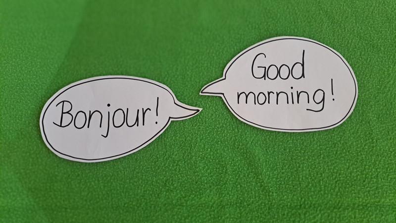 Gezeigt werden zwei Sprechblasen. Die linke Sprechblase enthält den französischen Gruß "Bonjour!" und die rechte Sprechblase den englischen Gruß "Good morning!".