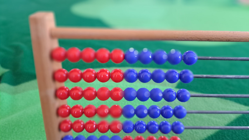 Auf dem Bild ist ein Abakus zu sehen, ein traditionelles Rechenhilfsmittel. Die Perlen sind in zwei Farben, rot und blau, angeordnet. Links befinden sich immer 5 rote Perlen und rechts 5 blaue Perlen.